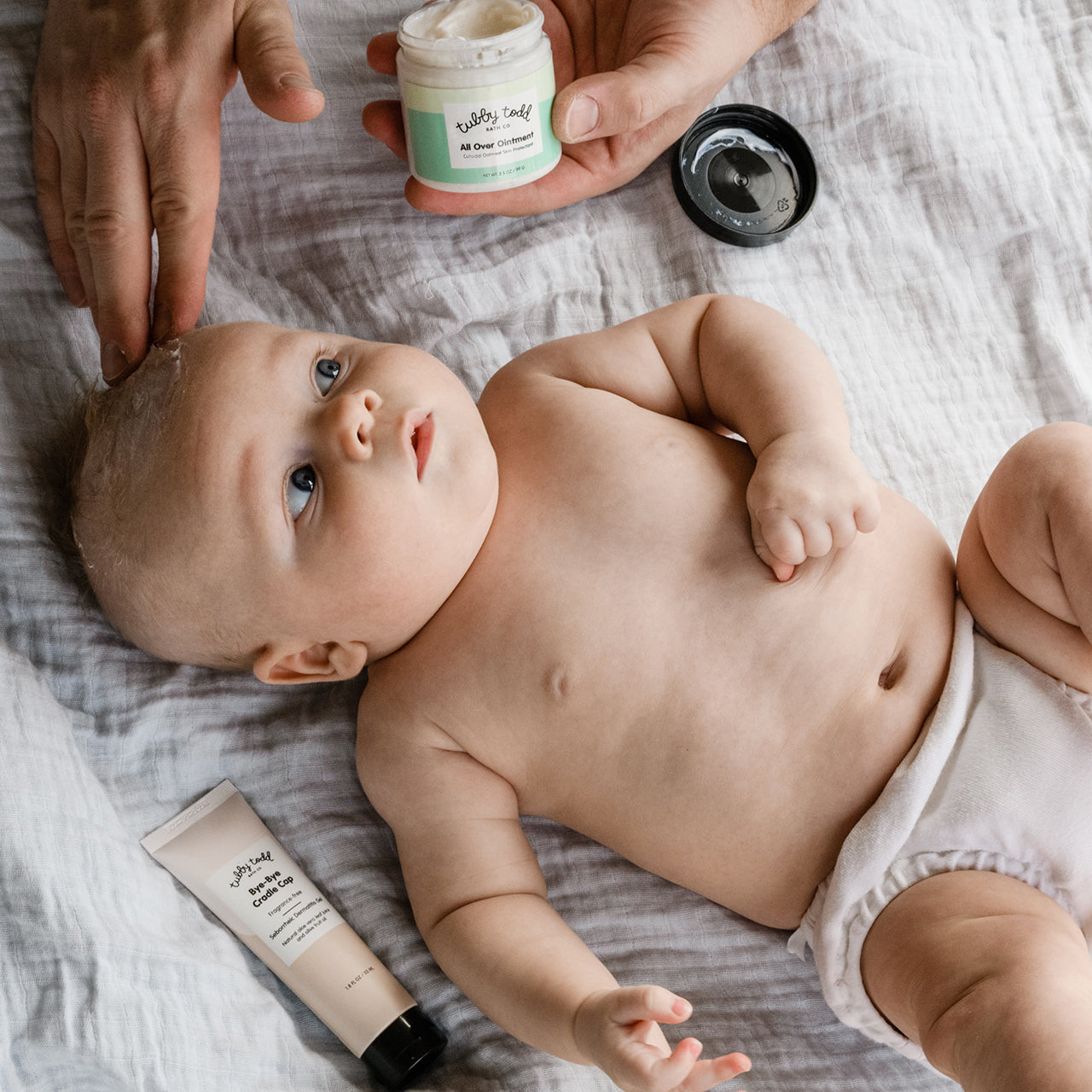 Dad applying All Over Ointment to baby's scalp on bed